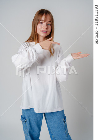 Young girl happily pointing against a neutral background while wearing a cozy white sweatshirt 119511393