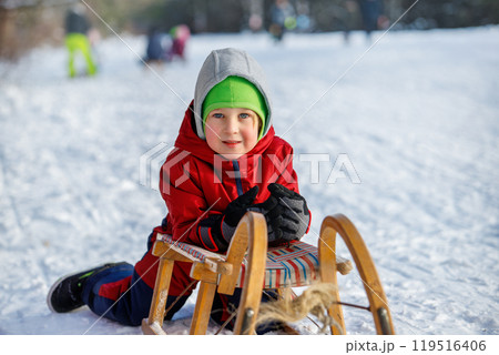 Happy Little boy kid portrait wear overall enjoy having fun sledding vintage wooden sledge on snowy hill in city park outdoors at winter holiday. Smiling child riding sport bob outside 119516406
