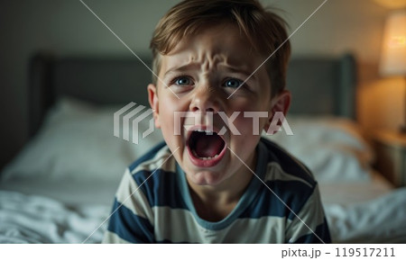 A young boy is sitting on a bed, crying loudly with his mouth open wide A young boy is sitting on a bed, crying loudly with his mouth open wide 119517211