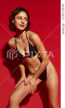 Fit young woman posing in black bikini, put her hands on leg, as if she applying sun-protective cream against vivid red background. Fit young woman posing in black bikini, put her hands on leg, as if she applying sun-protective cream against vivid red background. 119517676