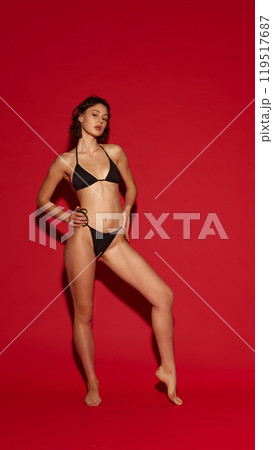 Young attractive woman poses gracefully in black bikini, showing her athletic body forms against vivid red background. Young attractive woman poses gracefully in black bikini, showing her athletic body forms against vivid red background. 119517687