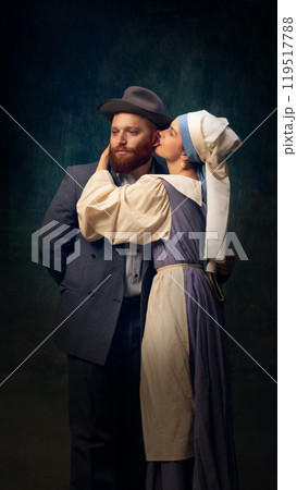 Flirty mood. Beautiful young woman in vintage dress and headscarf standing next to man in classical suit against dark vintage background. Flirty mood. Beautiful young woman in vintage dress and headscarf standing next to man in classical suit against dark vintage background. 119517788