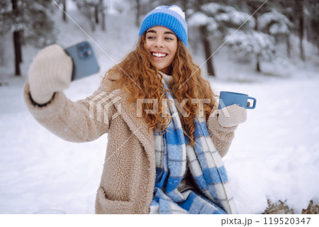Portrait of a beautiful woman in warm clothes takes selfie with a hot drink from a thermos in hands. 119520347