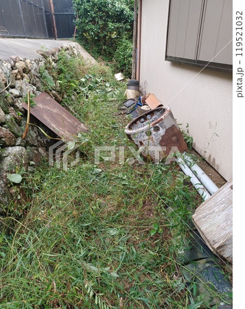 不用品の散乱する草刈り中の裏庭、左に隣家との境界の石垣と目隠し用の黒いフェンス 119521032