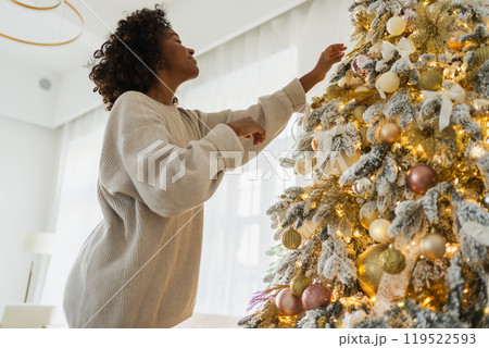 Merry Christmas. African American woman decorating Christmas tree. Happy girl near classical Christmas tree with white golden silver decorations ornament. Christmas eve at home time for celebration 119522593