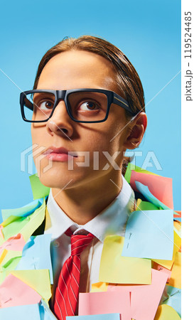 Thoughtful young man in glasses, wearing suit with red tie, standing fully covered in multicolored sticky notes against blue background. 119524485