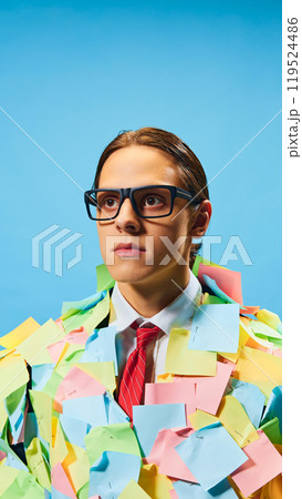 Creative portrait of young man in glasses, wearing suit with red tie, standing fully covered in multicolored sticky notes against blue background. 119524486
