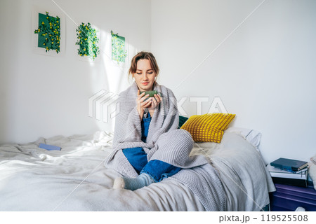 Woman freezes in wintertime. Young female student wearing warm woollen socks and wrapped into blanket, holding cup of hot drink while sitting on the bed at home. Keep warm. Bills for Cost of heating. Woman freezes in wintertime. Young female student wearing warm woollen socks and wrapped into blanket, holding cup of hot drink while sitting on the bed at home. Keep warm. Bills for Cost of heating. 119525508
