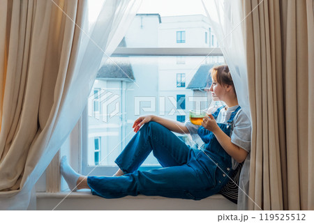 Free Time To Relax. Young woman looks out the window overlooking the city, sits on the windowsill at cozy home holds cup of hot tea drink. Happy calm female taking break for mental health wellbeing Free Time To Relax. Young woman looks out the window overlooking the city, sits on the windowsill at cozy home holds cup of hot tea drink. Happy calm female taking break for mental health wellbeing 119525512
