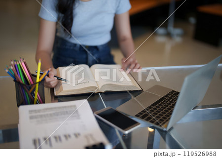 Asian teenage student woman reading a book to writing information from book and doing assignment homework after watching video online lesson on laptop and studying about education knowledge in cafe Asian teenage student woman reading a book to writing information from book and doing assignment homework after watching video online lesson on laptop and studying about education knowledge in cafe 119526988