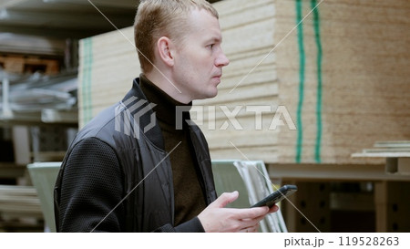 Man in a hardware store, choosing lumber, shelves with lumber. Man uses a smartphone to select plywood in a hardware store for a home construction or renovation project Man in a hardware store, choosing lumber, shelves with lumber. Man uses a smartphone to select plywood in a hardware store for a home construction or renovation project 119528263