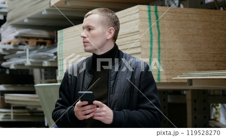 Contractor uses his smartphone to search for building material prices while in the hardware store aisle. A man in a hardware store, choosing lumber, shelves with lumber. 119528270