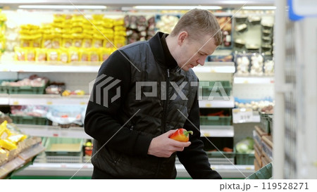 Man thoughtfully chooses fresh produce at the supermarket, examining a bright bell pepper and checking the label for freshness. Man choosing fruits in supermarket, shelf with sweet peppers 119528271