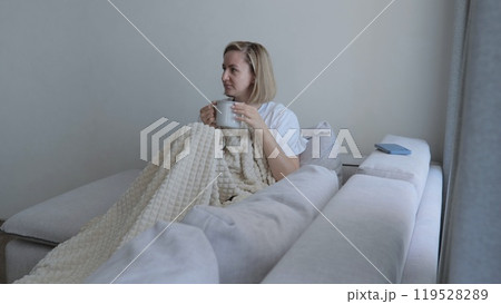 Young woman covered with a warm blanket drinks tea at home. A young woman drinks hot tea while lying on the sofa. A woman on the sofa, covered with a blanket, drinks a hot drink. 119528289
