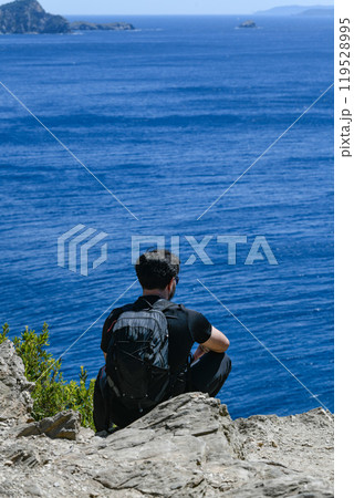 Young sporty man with backpack sitting on the top of rock and looking at the seashore and mountains 119528995