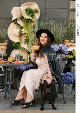 A girl is sitting in a summer cafe with her French bulldog and drinking coffee with a cake A girl is sitting in a summer cafe with her French bulldog and drinking coffee with a cake 119530251