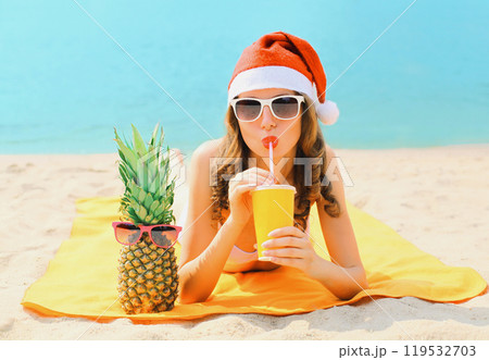 Christmas portrait woman in red santa hat with pineapple drinking juice lying on beach on sea Christmas portrait woman in red santa hat with pineapple drinking juice lying on beach on sea 119532703