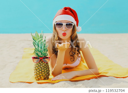 Christmas portrait woman in red santa hat with pineapple lying on beach blowing kiss on sea Christmas portrait woman in red santa hat with pineapple lying on beach blowing kiss on sea 119532704