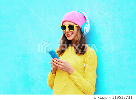 Happy smiling young woman listening to music with headphones holding phone on blue background Happy smiling young woman listening to music with headphones holding phone on blue background 119532731
