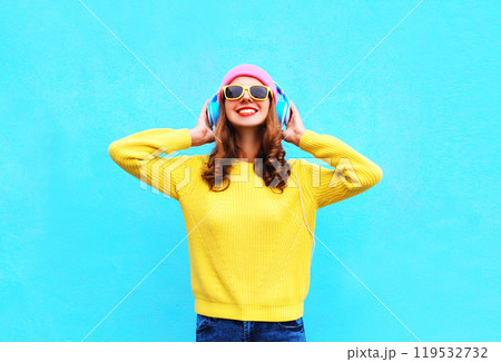 Happy young woman listening to music with headphones in colorful yellow sweater on blue background Happy young woman listening to music with headphones in colorful yellow sweater on blue background 119532732