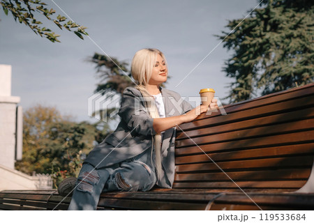 A blonde woman sits on a bench with a cup of coffee in her hand. She is wearing a gray jacket and jeans. The bench is wooden and has a curved back. The scene is peaceful and relaxing. A blonde woman sits on a bench with a cup of coffee in her hand. She is wearing a gray jacket and jeans. The bench is wooden and has a curved back. The scene is peaceful and relaxing. 119533684
