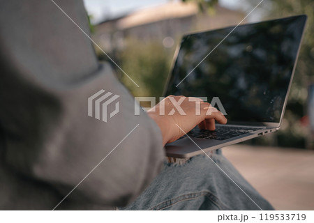 A person is sitting on a bench with a laptop open in front of them. They are typing on the keyboard, possibly working or browsing the internet. Concept of productivity and focus. A person is sitting on a bench with a laptop open in front of them. They are typing on the keyboard, possibly working or browsing the internet. Concept of productivity and focus. 119533719