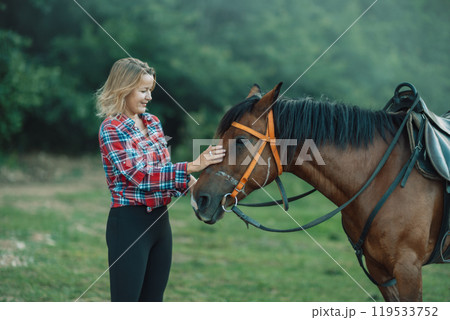 Happy blonde with horse in forest. Woman and a horse walking through the field during the day. Dressed in a plaid shirt and black leggings. 119533752