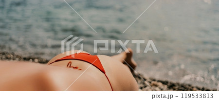 A woman sunbathes on the beach, lying on her stomach in a red swimsuit against the sea backdrop. A woman sunbathes on the beach, lying on her stomach in a red swimsuit against the sea backdrop. 119533813