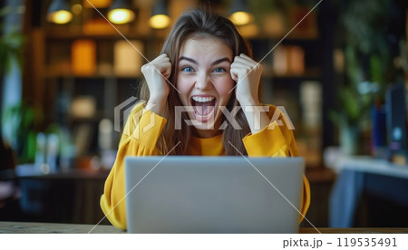Successful Young Business Woman In Her 20s, Happily Working On Laptop At Wooden Office Desk In Casual Yellow Shirt. Celebrating Career Achievement. 119535491