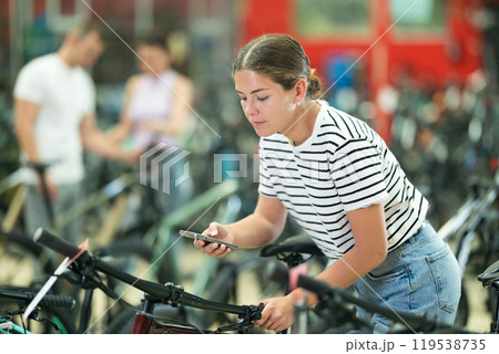 Young girl using smartphone for examining quality seat and choosing bike in bicycle sports store Young girl using smartphone for examining quality seat and choosing bike in bicycle sports store 119538735