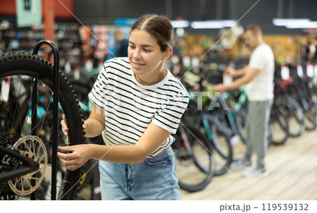 Glad young girl selects bicycle in sporting goods store, checking wheel quality 119539132