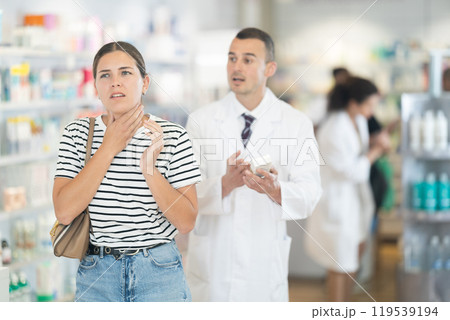 Young girl feeling pain standing with her back to male pharmacist offering remedy Young girl feeling pain standing with her back to male pharmacist offering remedy 119539194