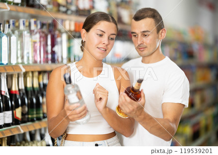 Man with wife near display of alcohol drinks in hypermarket doubt choice. Man with wife near display of alcohol drinks in hypermarket doubt choice. 119539201