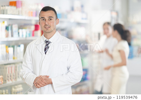 Positive male pharmacist in medical uniform posing while working in pharmacy Positive male pharmacist in medical uniform posing while working in pharmacy 119539476
