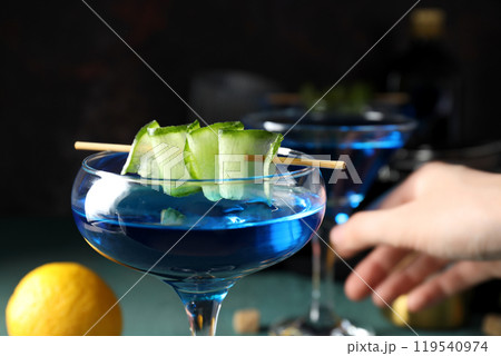 Blue cocktail with cucumber slices in glasses and hand on dark background, close up 119540974