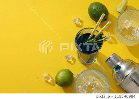 Glasses with ice cubes and blue cocktail, limes and shaker on yellow background, space for text Glasses with ice cubes and blue cocktail, limes and shaker on yellow background, space for text 119540984