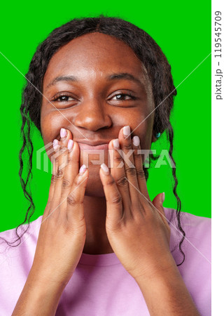 A young woman expresses joy while smiling and touching her face against a bright green background A young woman expresses joy while smiling and touching her face against a bright green background 119545709