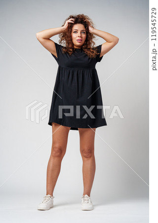Smiling woman with curly hair poses confidently against a plain background while holding her hands behind her head 119545729