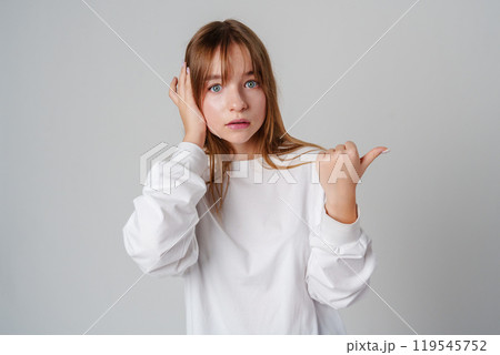 Young girl happily pointing against a neutral background while wearing a cozy white sweatshirt 119545752
