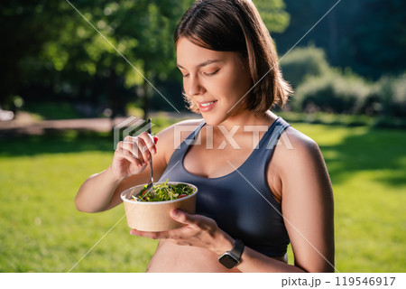 Pregnant woman holding plate with salad eating after yoga exercising sitting on green grass Pregnant woman holding plate with salad eating after yoga exercising sitting on green grass 119546917