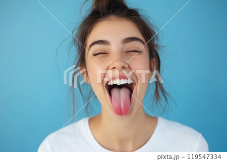 Young woman playfully sticking her tongue out against a vibrant blue background while wearing a 119547334