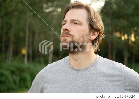 Portrait of handsome young man in gray t-shirt outdoors. Male with long hair and beard feel happy, calm and serious on Green nature sunset background. Man looking away into distance. Diversity People 119547624