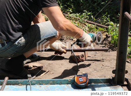partially visible man with grinder in the process of sawing off part of iron pipe, work with angle saw and iron materials 119549372