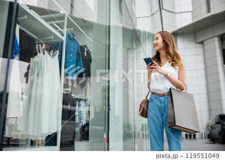 Beautiful woman with shopping bags, browsing her cell phone, standing by a fashion store display. She is enjoying the convenience of modern technology and the pleasure of consumerism. Beautiful woman with shopping bags, browsing her cell phone, standing by a fashion store display. She is enjoying the convenience of modern technology and the pleasure of consumerism. 119551049
