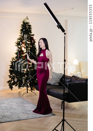 A beautiful brunette woman with makeup in a red dress celebrates the new year and sings songs in a decorated festive interior of a room with a Christmas tree on a holiday at home 119555355