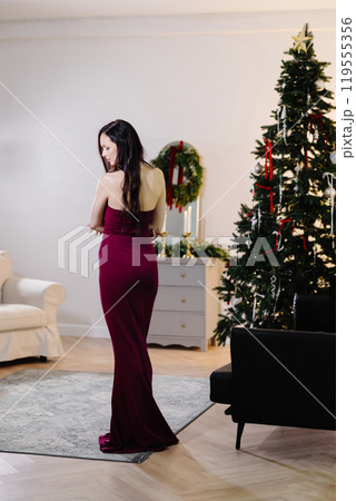 A beautiful brunette woman with makeup in a red dress celebrates the new year and sings songs in a decorated festive interior of a room with a Christmas tree on a holiday at home 119555356
