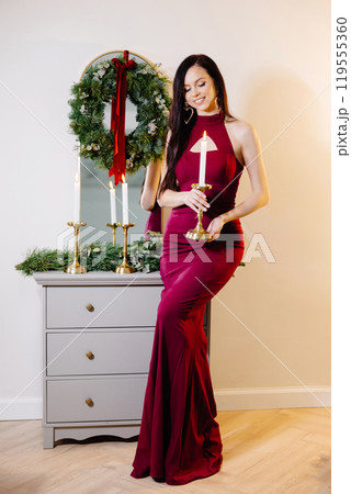 A beautiful brunette woman with makeup in a red dress celebrates the new year in a decorated festive interior of a room with a Christmas tree on a holiday at home 119555360