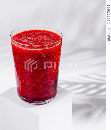 Glass of red berry smoothie sitting on white cube with tropical plant shadow Glass of red berry smoothie sitting on white cube with tropical plant shadow 119555691