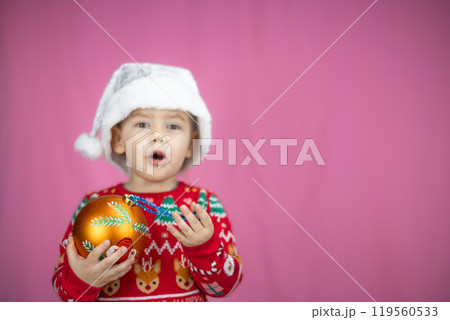 Cute child in a festive sweater and Santa hat holds a Christmas decoration on a pink background. The child is happy about the New Year and Christmas. Suitable for holiday projects. 119560533