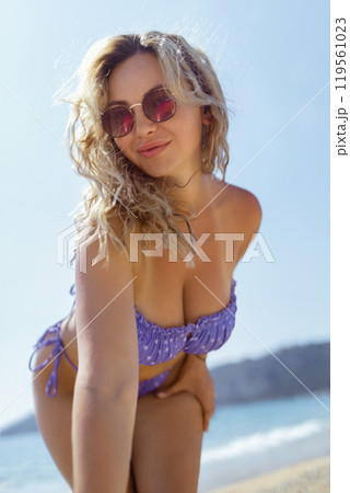 A beautiful girl in sunglasses on the beach looks at the camera, leaning towards it. Sexy blonde in a swimsuit on the seashore A beautiful girl in sunglasses on the beach looks at the camera, leaning towards it. Sexy blonde in a swimsuit on the seashore 119561023
